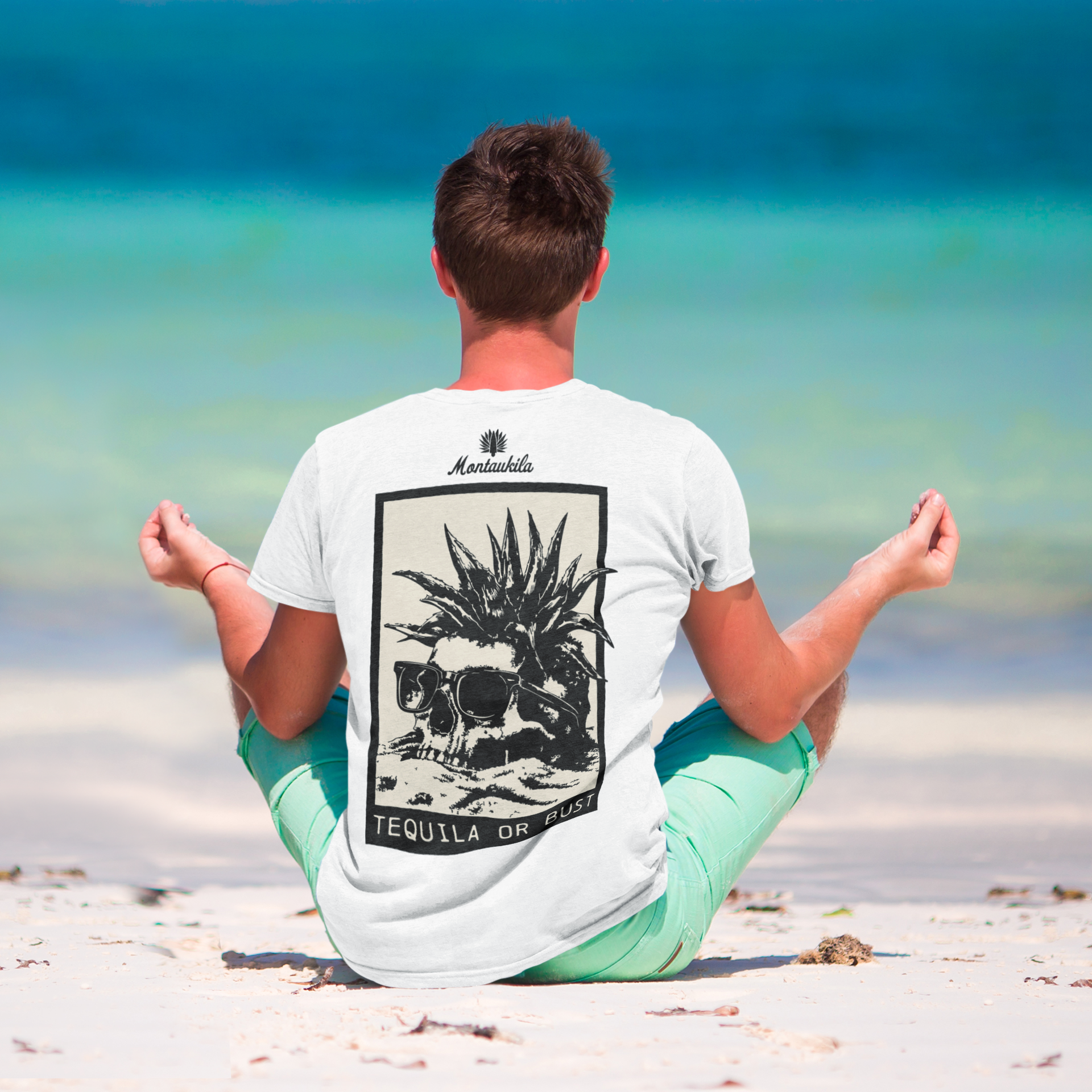 Person sitting on a beach wearing a white t-shirt with a montaukila design